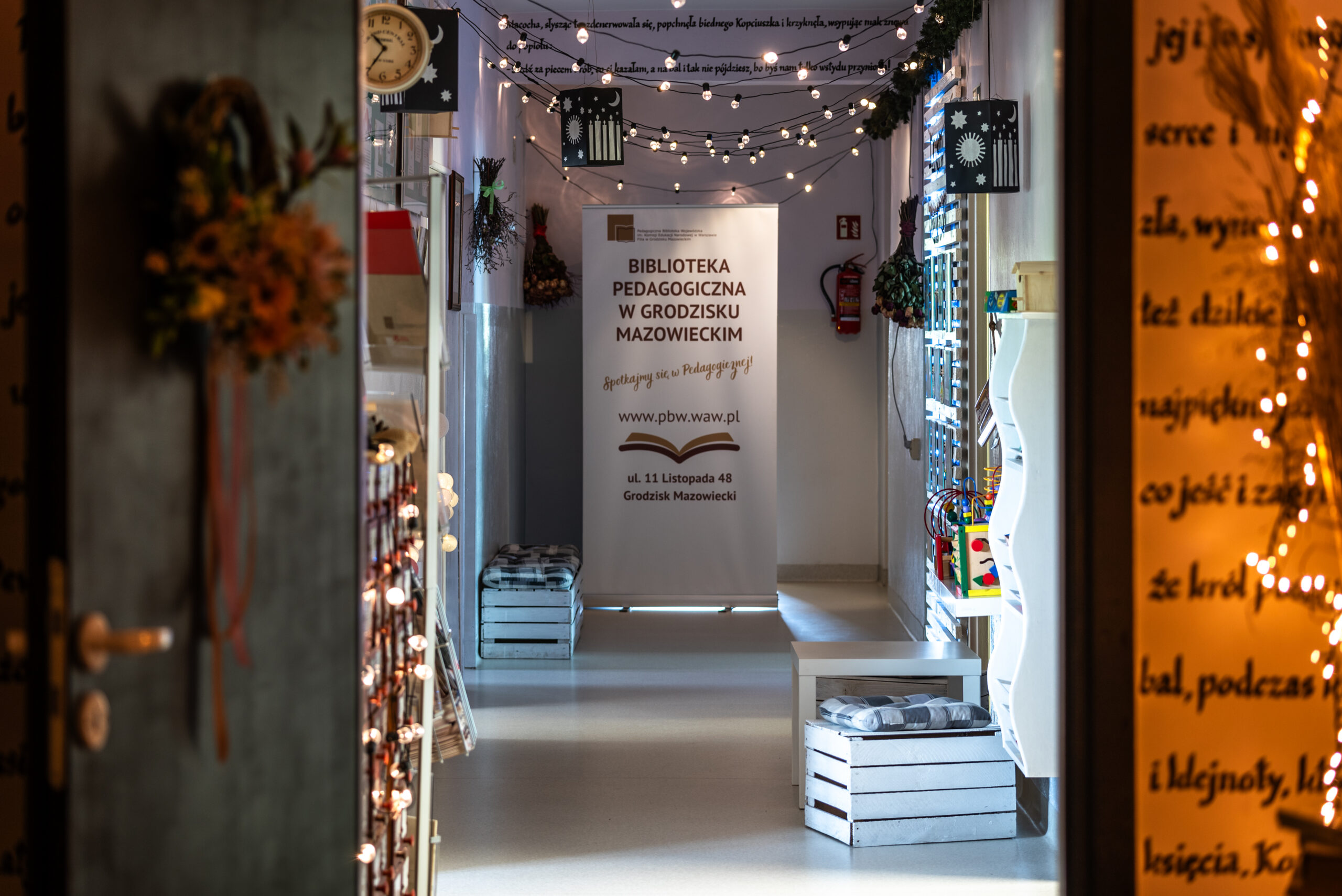 Korytarz prowadzący do Biblioteki Pedagogicznej w Grodzisku Mazowieckim. W centralnym punkcie stoi biały banner z logo i adresem placówki. Korytarz ozdobiony jest nastrojowymi sznurami żarówek podwieszonymi pod sufitem, suszonymi kwiatami na ścianach oraz dekoracyjnymi lampionami. Po bokach znajdują się niskie białe regały z zabawkami edukacyjnymi i siedziska wykonane z drewnianych skrzyń z poduszkami w kratę